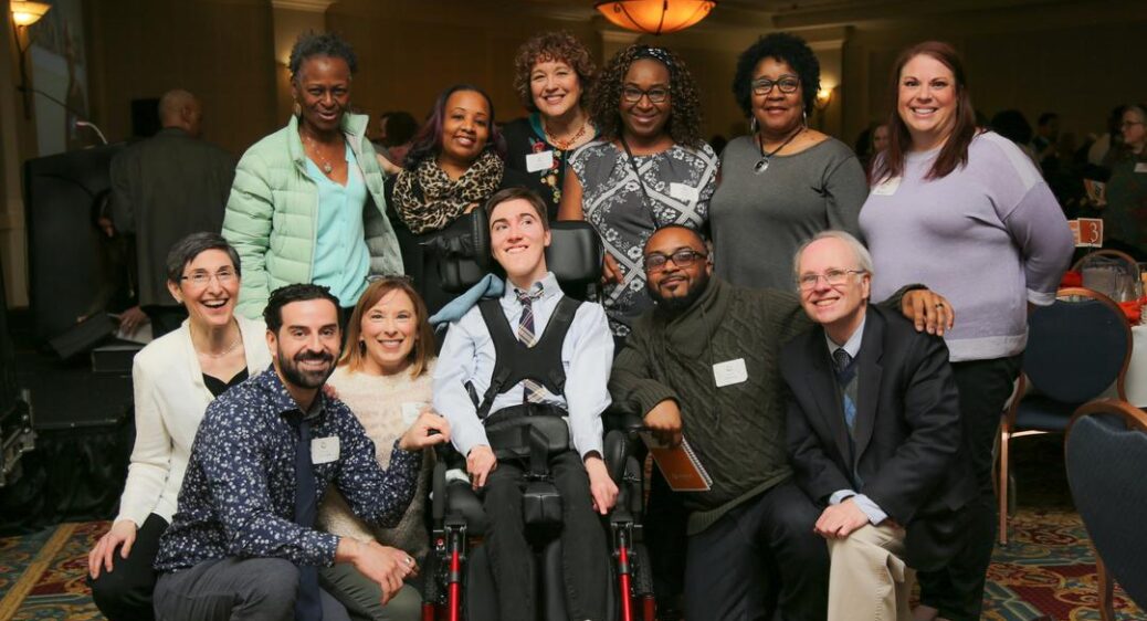 Smiling young man in a wheelchair with a team of family and providers smiling on all sides of him