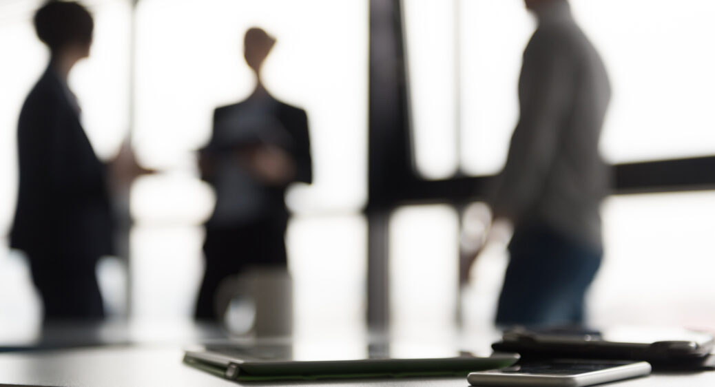 Close up of smart phone and tablet computer at office meeting room. 3 blurred silhouettes of people interacting in background.
