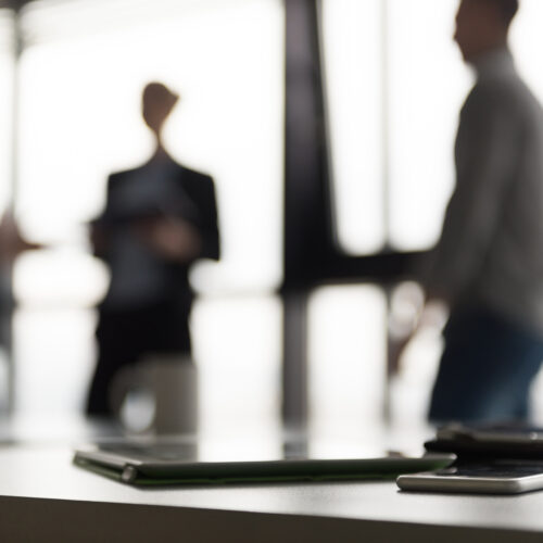 Close up of smart phone and tablet computer at office meeting room. 3 blurred silhouettes of people interacting in background.